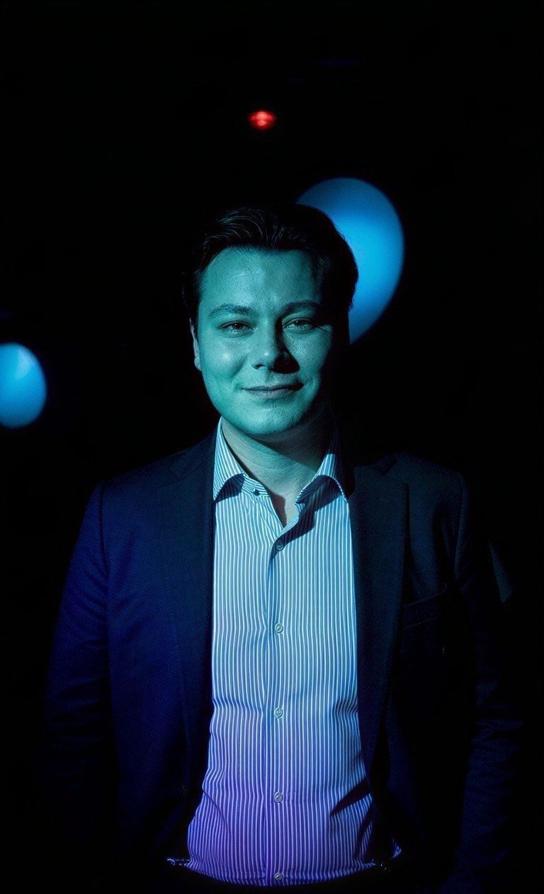 Portrait de Clément Perrin en veste sombre et chemise rayée, éclairé d’une lumière cyan